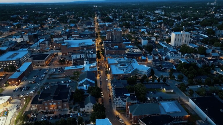 Aerial view of a small city with a mix of modern and historic buildings, a church with a tall steeple, tree-lined streets, and the evening sky.